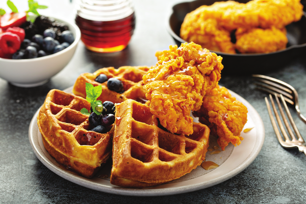 A plate of golden-brown waffles topped with fresh blueberries and a sprig of mint, drizzled with syrup, accompanied by crispy fried chicken. In the background, there's a bowl of assorted berries, a jar of syrup, and more fried chicken on a dark skillet. The meal is served on a dark surface, with a pair of forks resting beside the plate, signaling a ready-to-enjoy hearty meal.