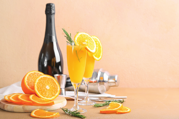 Two flutes of mimosa cocktail adorned with orange slices and rosemary sprigs, standing next to a bottle of champagne and a cocktail shaker on a light-colored table. Slices of fresh orange are arranged on a wooden cutting board in the foreground, with a white napkin and bar utensils nearby. The warm, pastel background creates a cheerful and inviting setting for a festive brunch or celebration.