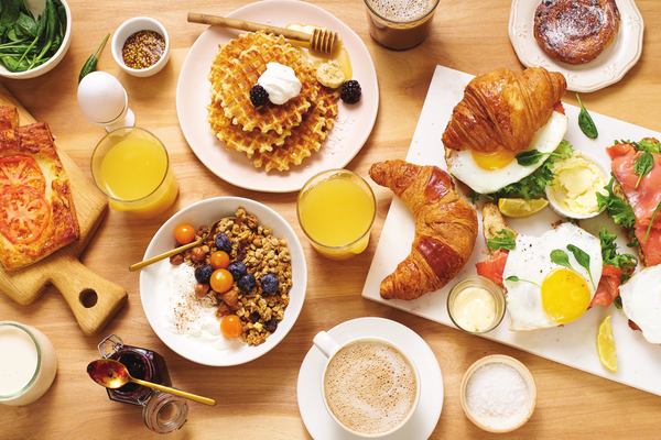 A breakfast table laden with an array of dishes: a stack of golden waffles topped with whipped cream and blackberries, a bowl of granola with blueberries and yogurt, freshly baked croissants, one topped with smoked salmon and a fried egg, alongside a rolled omelet with tomato slices. The scene includes glasses of orange juice, cups of coffee, a small bowl of spinach, honey with a dipper, and a jar of jam. The table presents a delightful and inviting spread of morning favorites.